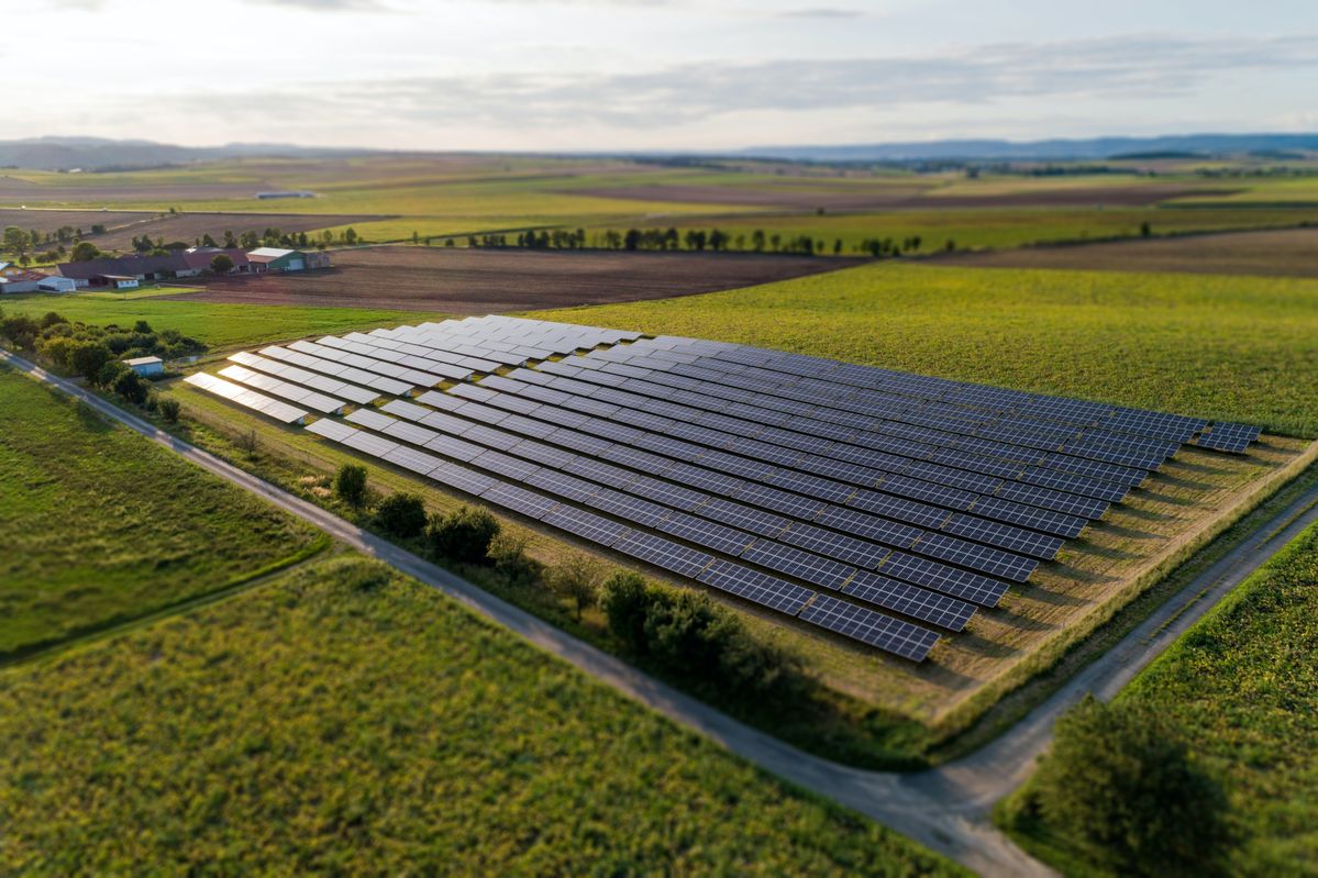 Aerial view of a large solar farm installed in an open green field with farmland and roads around it.