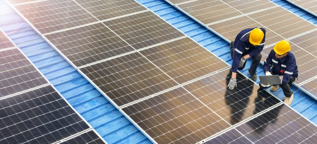 Two technicians inspecting rooftop solar panels on a metal roof while using a laptop during installation.