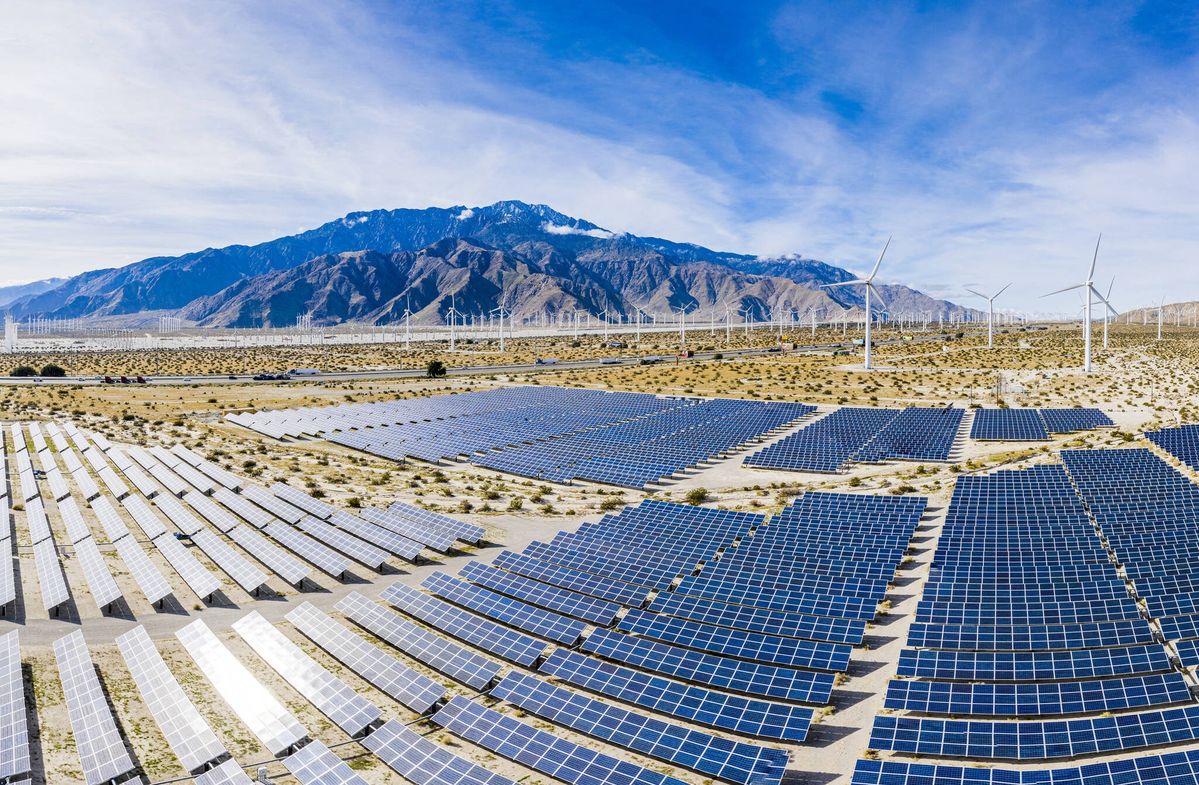 A large solar farm with rows of solar panels and wind turbines installed in a dry open landscape with mountains in the background.