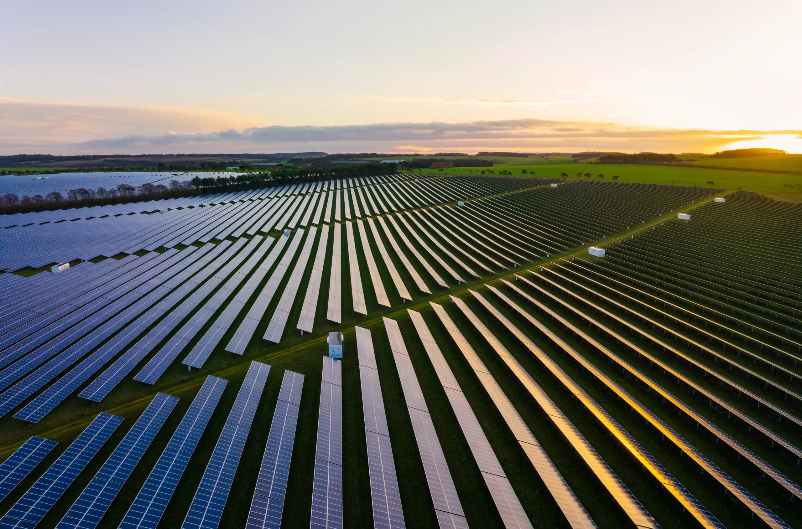 A wide aerial view of a large solar farm with rows of solar panels reflecting sunlight during sunset.
